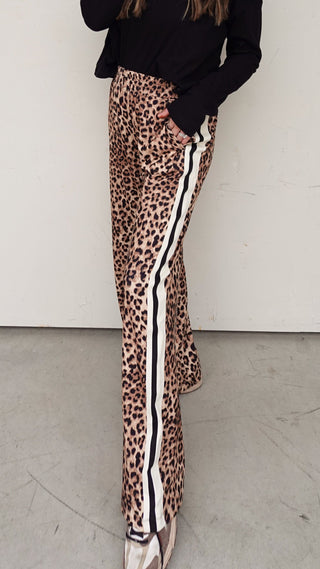 Woman wearing a black sweater and leopard print pants with white stripes against a white wall.