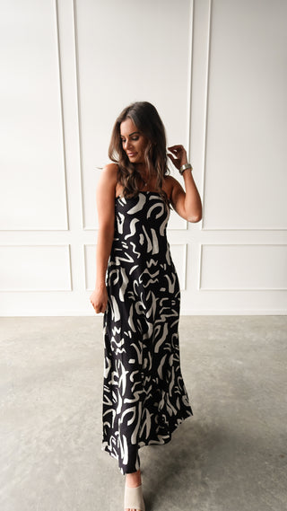 Model wearing a black strapless maxi dress with cream abstract print, standing in soft light in front of a white paneled wall.