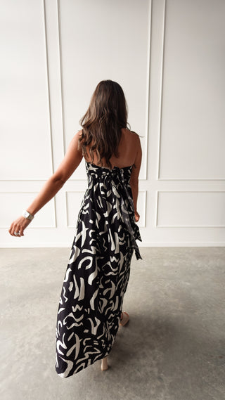 Model wearing a black strapless maxi dress with cream abstract print, standing in soft light in front of a white paneled wall.