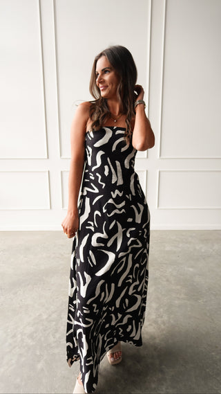 Model wearing a black strapless maxi dress with cream abstract print, standing in soft light in front of a white paneled wall.